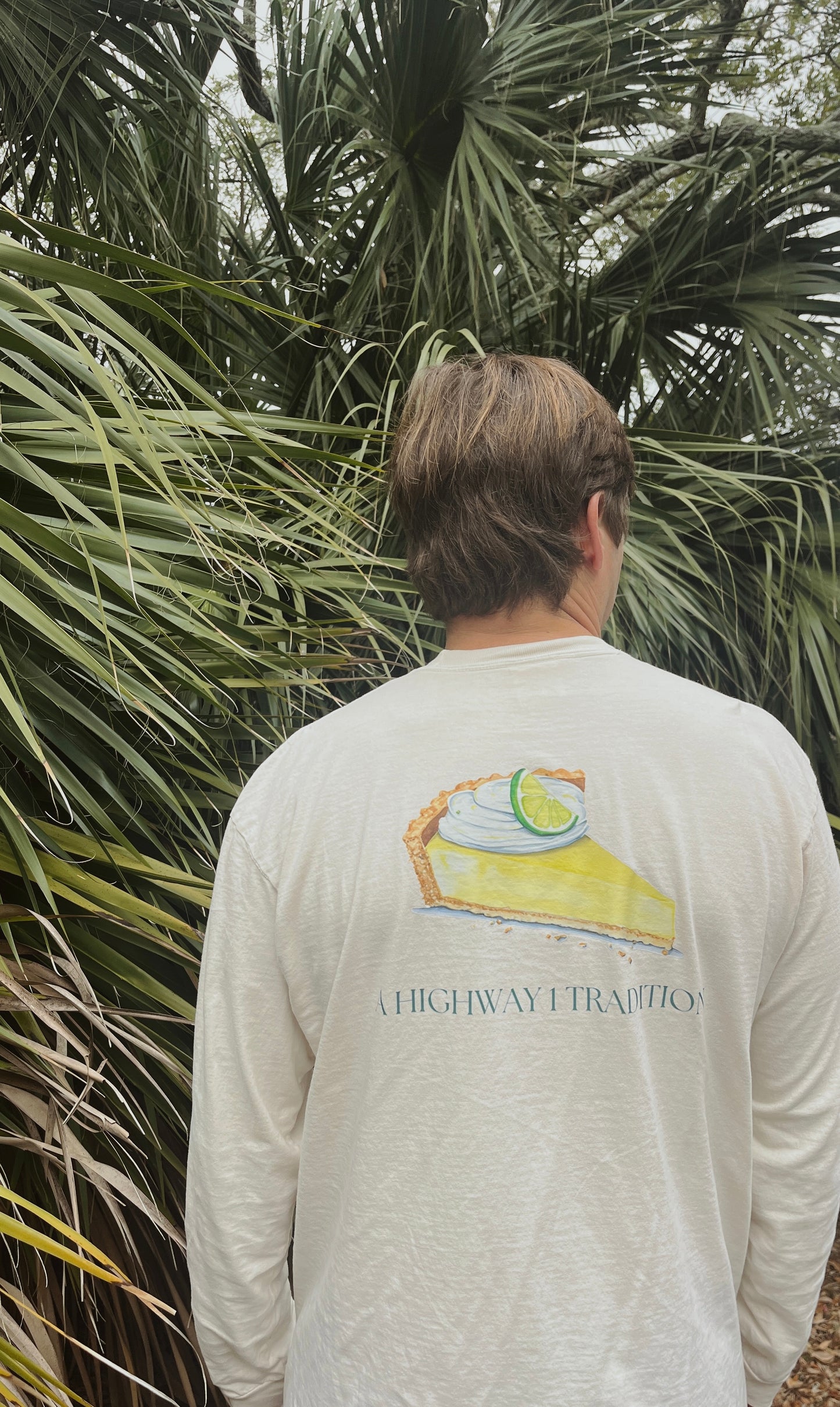 Person wearing a long-sleeve shirt with a pie graphic and 'Highway 1' text, standing in front of palm trees.