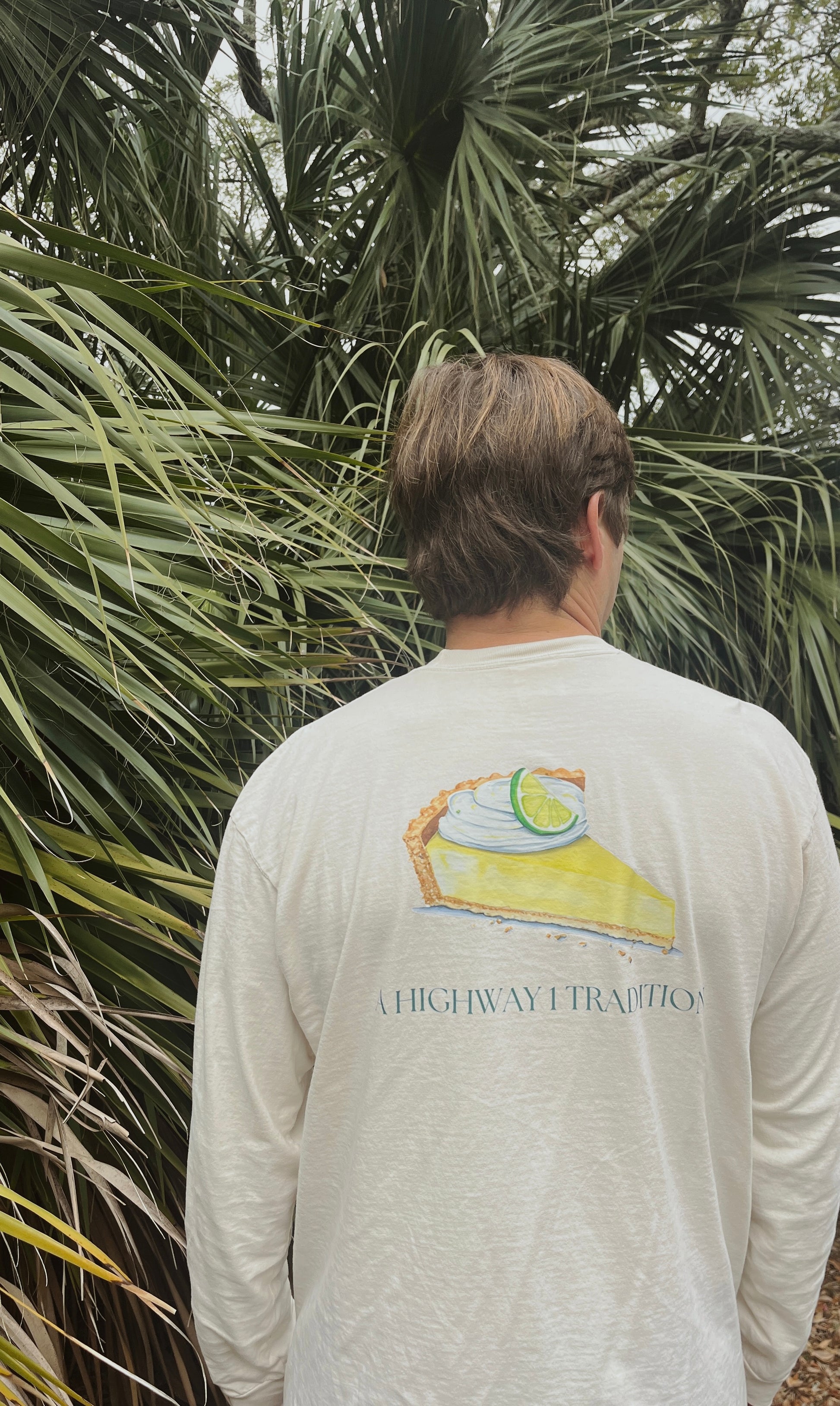 Person wearing a long-sleeve shirt with a pie graphic and 'Highway 1' text, standing in front of palm trees.