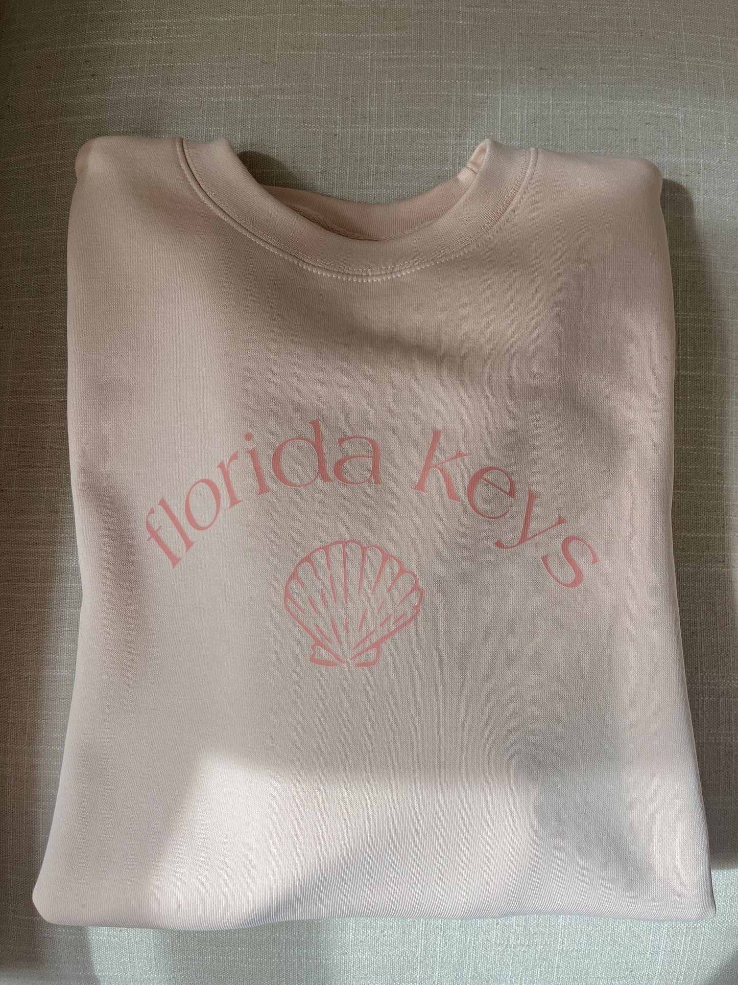 Folded pink sweatshirt with 'Florida Keys' and a shell design on a textured surface.
